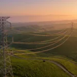 Vista aerea de torres e linhas de transmissao de alta tensao cruzando colinas verdes ao entardecer