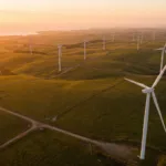 Vista aerea de um parque eolico com turbinas brancas em colinas verdes ao entardecer