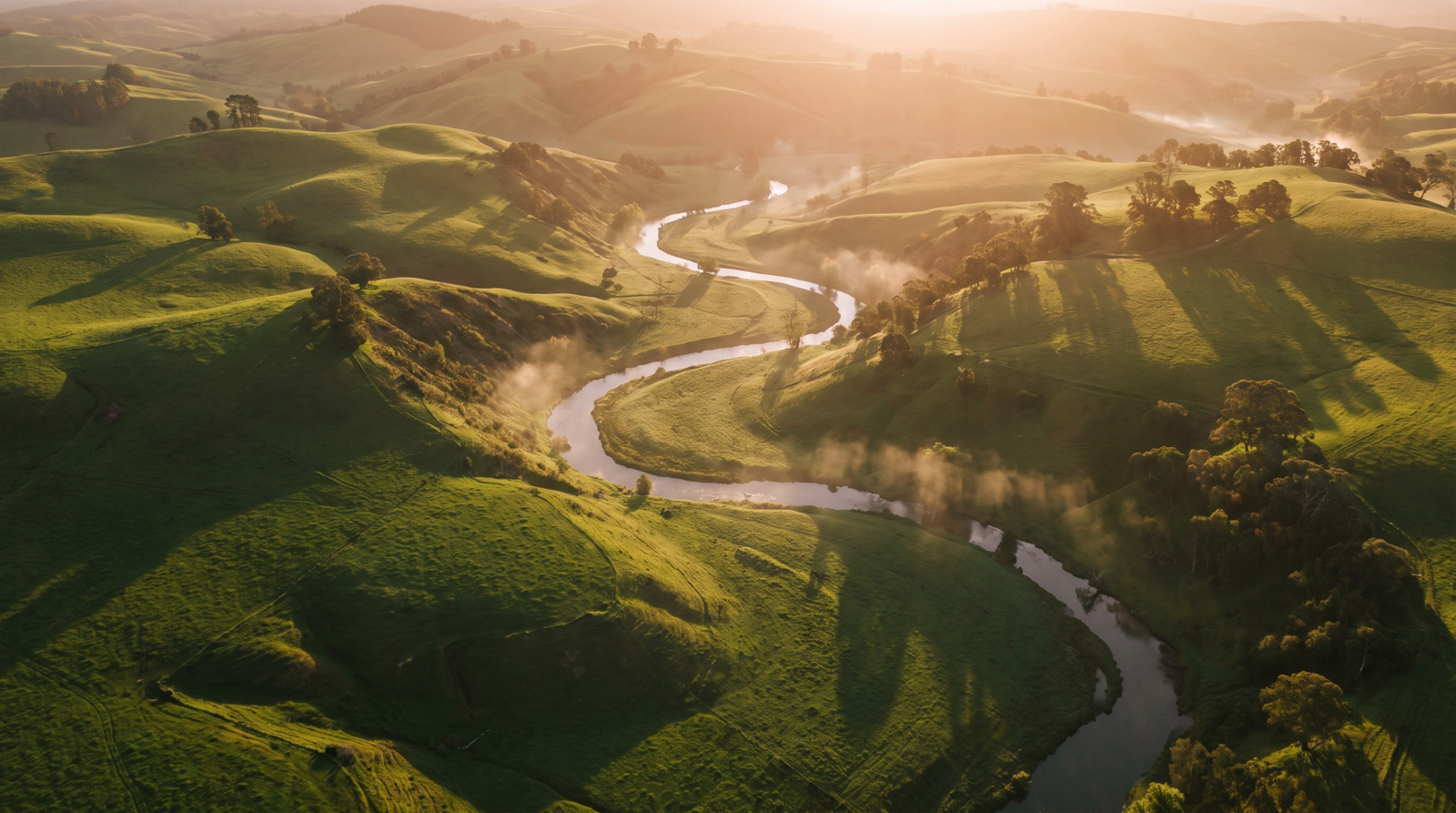 Foto aerea de uma paisagem na golden hour com sombras longas sobre morros verdes e um rio