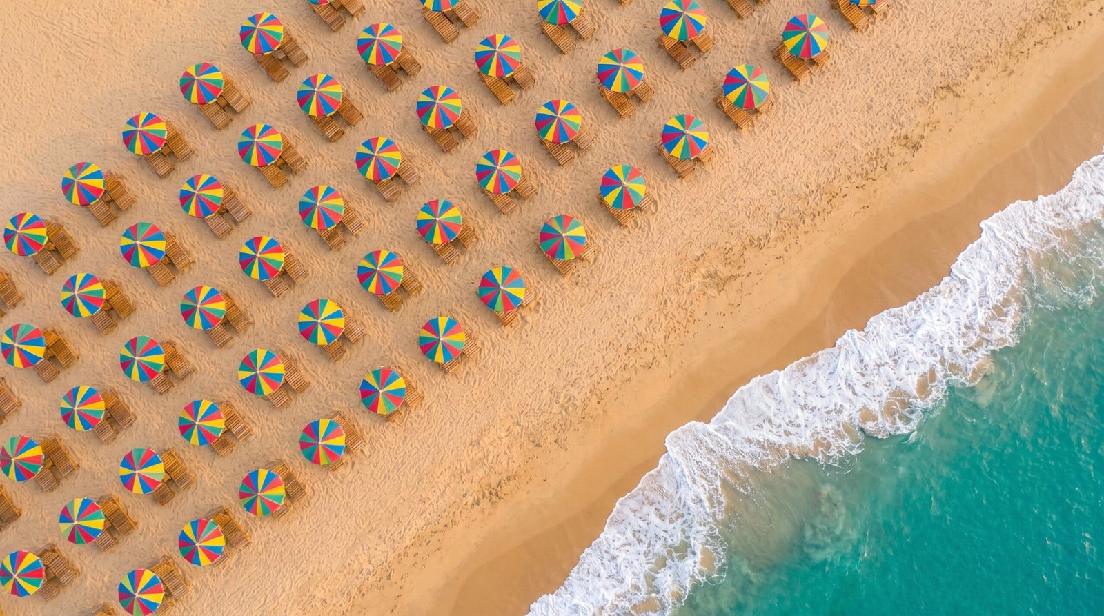 Vista top-down de uma praia com guarda-sois coloridos e a linha diagonal da espuma das ondas