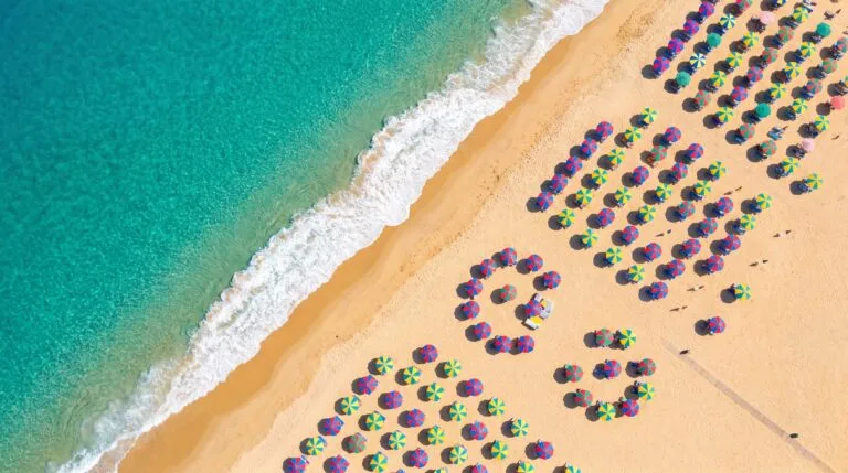 Foto aerea top-down de praia com mar turquesa, areia dourada e guarda-sois coloridos em padroes graficos