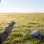 Piloto confere o celular ao amanhecer com o drone pousado na grama, pronto para decolar