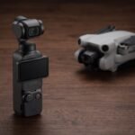 Câmera DJI Osmo Pocket 3 em pé sobre mesa escura com drone desfocado ao fundo