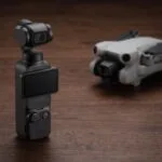 Câmera DJI Osmo Pocket 3 em pé sobre mesa escura com drone desfocado ao fundo