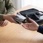 Duas pessoas trocando um drone dobrado sobre a mesa, com maleta e celular ao lado