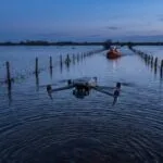 Drone com câmera térmica sobre estrada alagada, com bote de resgate ao fundo