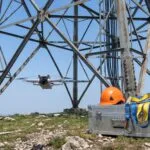 Drone pairando na base de uma torre com capacete e cinto de segurança ao lado