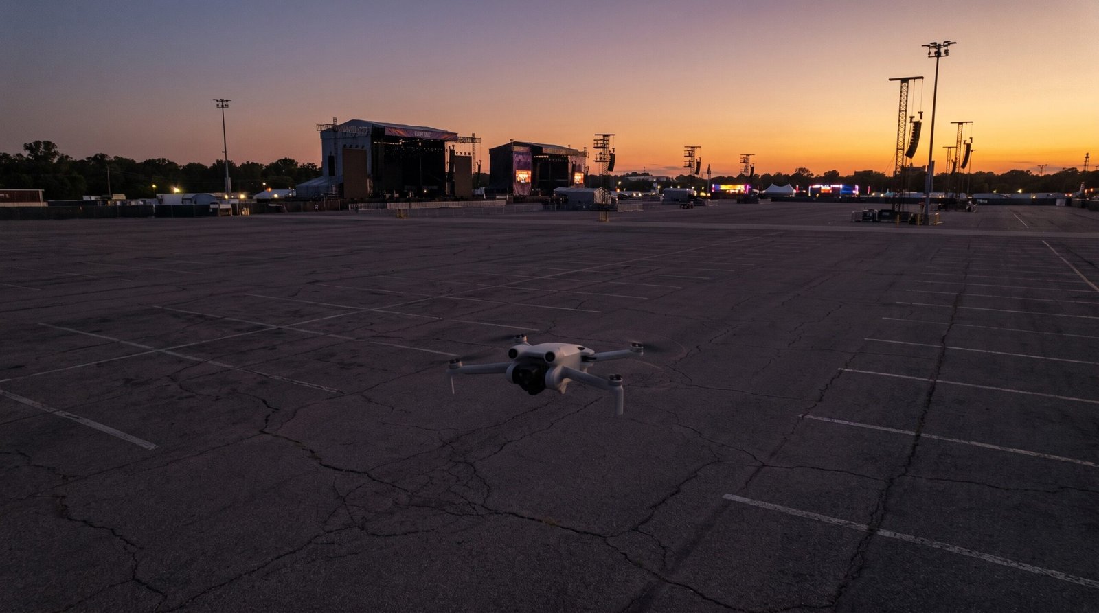 Drone pequeno pairando sobre uma área vazia, com as estruturas de um festival ao longe