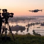 Equipe de TV filmando de um morro com drone sobre área alagada ao amanhecer