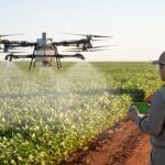 Drone agrícola DJI Agras T50 pulverizando lavoura de soja ao amanhecer, com o piloto na beira do talhão