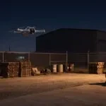 Drone pairando à noite sobre pátio industrial iluminado