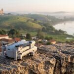 Drone compacto da linha DJI Mini dobrado sobre um muro de pedra, com uma cidade do Vale do Paraíba e o rio ao fundo ao nascer do sol