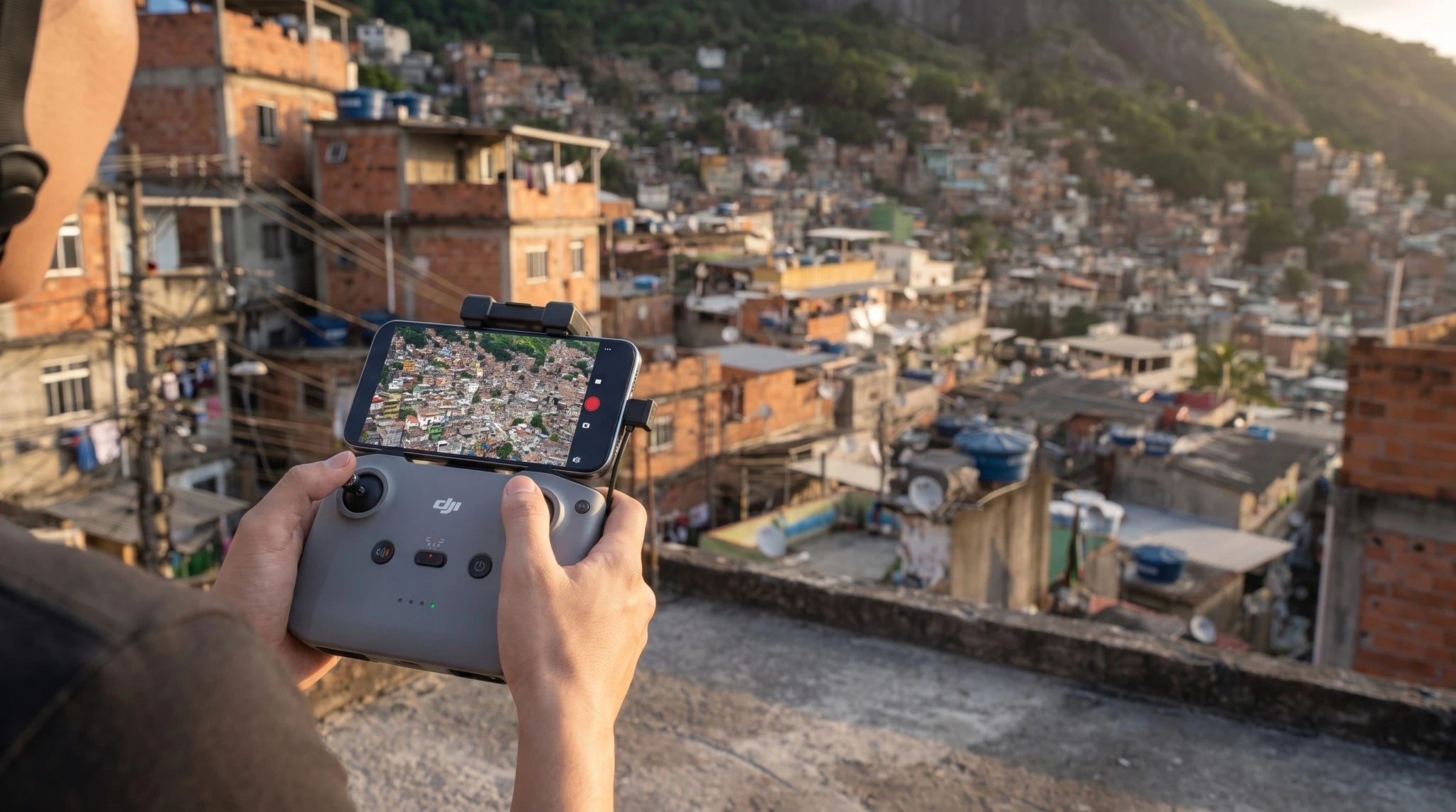 Mãos do piloto no controle DJI com celular acoplado e o morro da Rocinha desfocado ao fundo