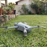 Drone compacto da linha DJI Mini caído na grama de um quintal ao lado de um muro baixo, de manhã