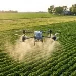 Drone agrícola DJI Agras T50 pulverizando soja com uma casa de fazenda ao fundo, longe da área aplicada