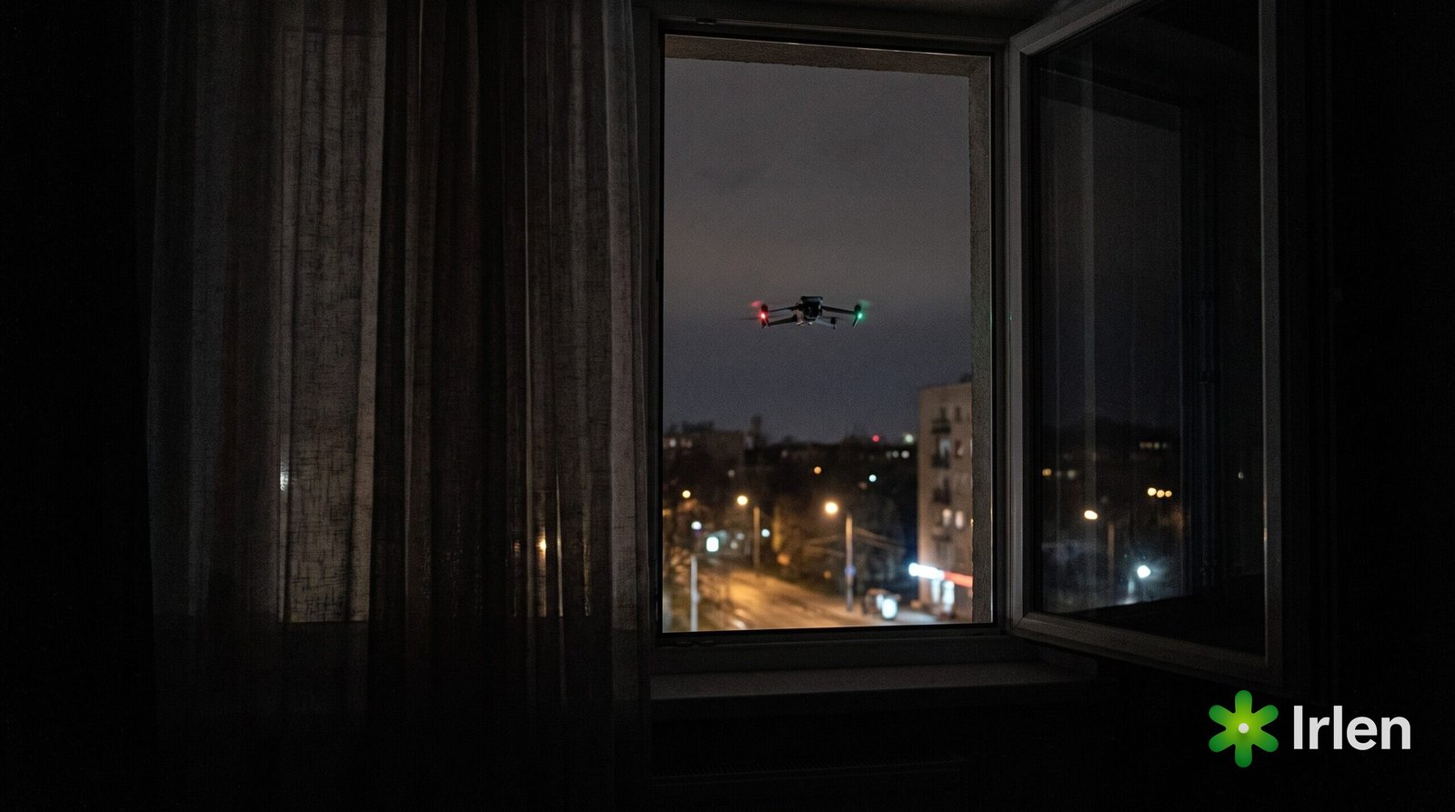 Vista de uma janela à noite com um drone DJI da família Mavic 3 parado ao longe sobre a rua