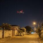 Drone DJI da família Mavic 3 parado no alto, à noite, sobre uma rua residencial com casas e muros