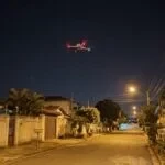 Drone DJI da família Mavic 3 parado no alto, à noite, sobre uma rua residencial com casas e muros