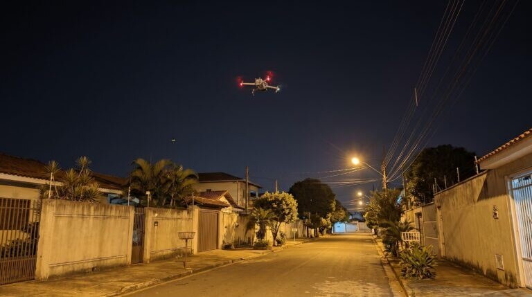 Drone DJI da família Mavic 3 parado no alto, à noite, sobre uma rua residencial com casas e muros